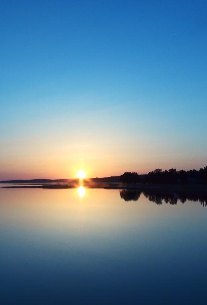 Tyyni suomalainen järvimaisema, jossa aurinko on nousemassa horisontista vastarannalta puiden takaa.